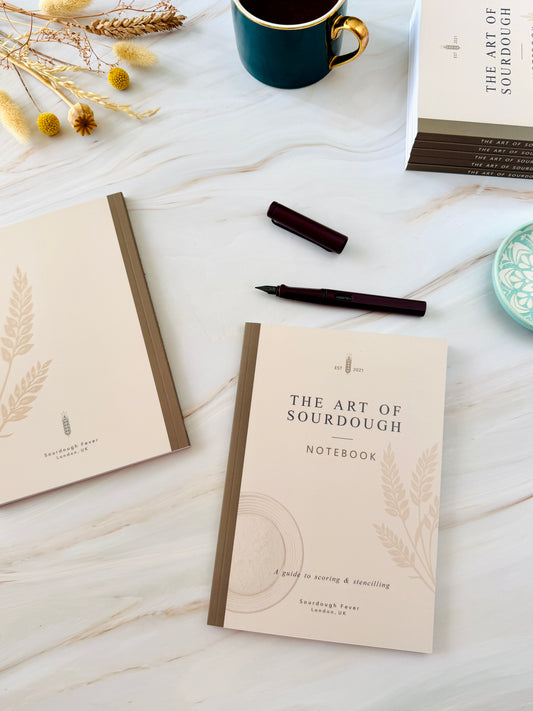 A styled flat-lay of a notebook titled 'The Art of Sourdough' on a white marble surface, alongside a pen, a coffee mug, dried flowers, and another copy of the notebook.