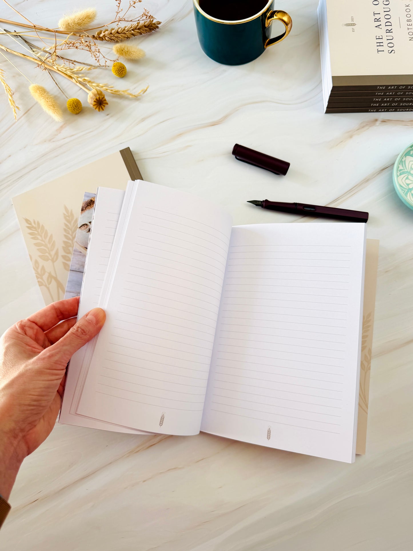 A hand holding an open lined notebook on a light marble desk, surrounded by a dark teal mug, dried flowers, a pen, and a book.