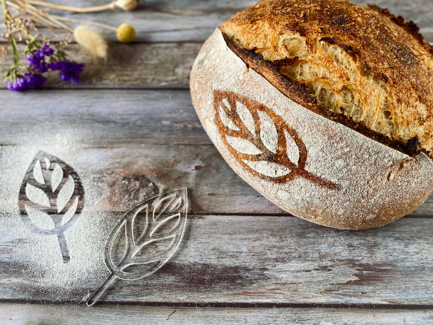 Loaf of bread with leaf design on a wooden surface, accompanied by a stenciled leaf and flour.