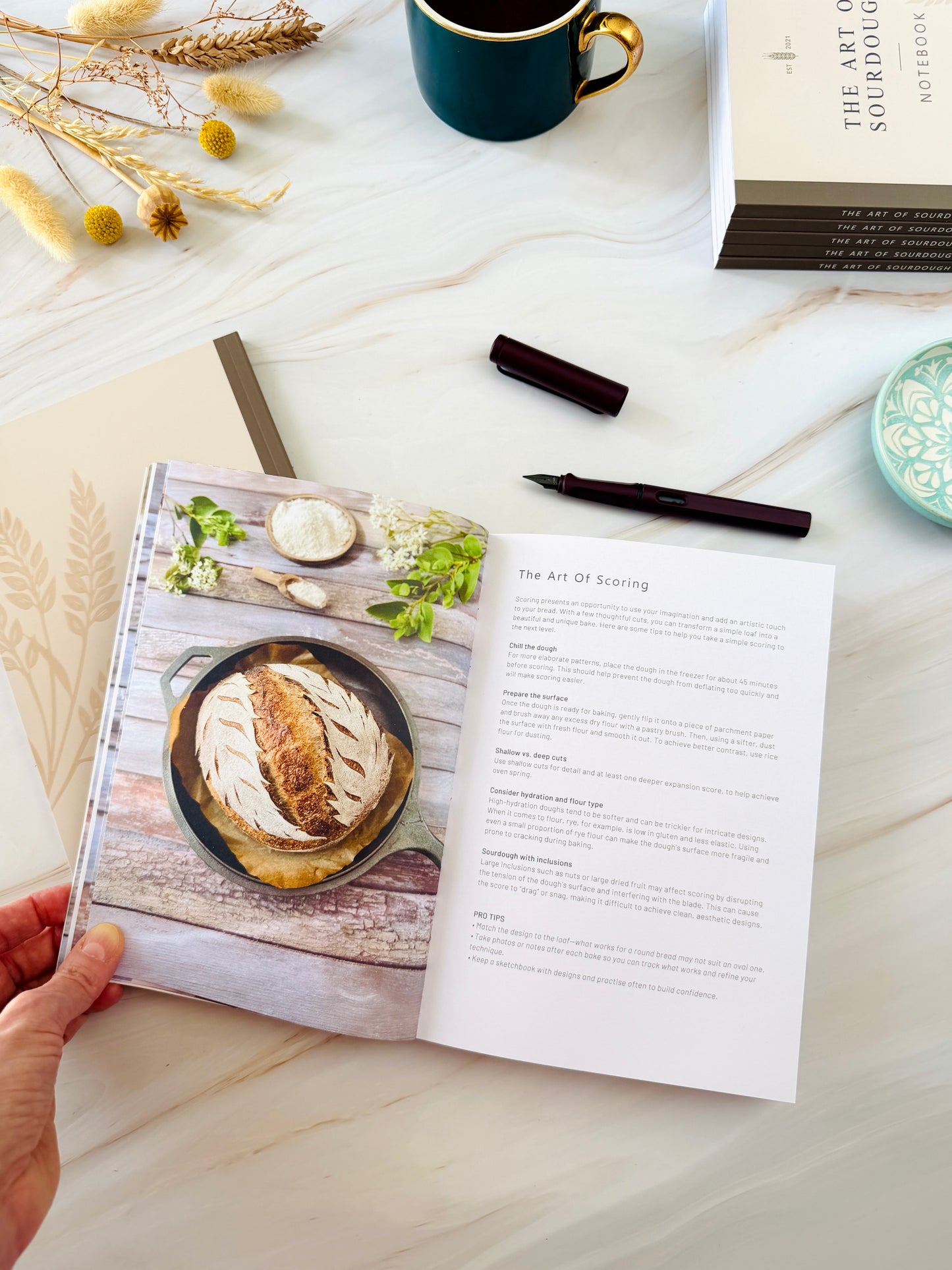 A hand holds an open cookbook on a light marble surface, showing a food photography page with a roasted fish in a skillet and a recipe page. A dark teal mug, dried flowers, a pen, and a stack of notebooks are arranged around it.