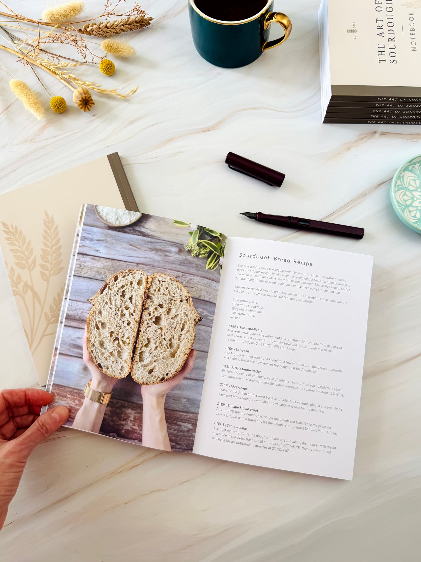 A hand holds an open recipe book showing a sourdough bread recipe page, with a printed photo of hands holding sliced bread on the left page. The scene is styled on a light marble surface with a dark mug, dried flowers, a pen, and other small props.