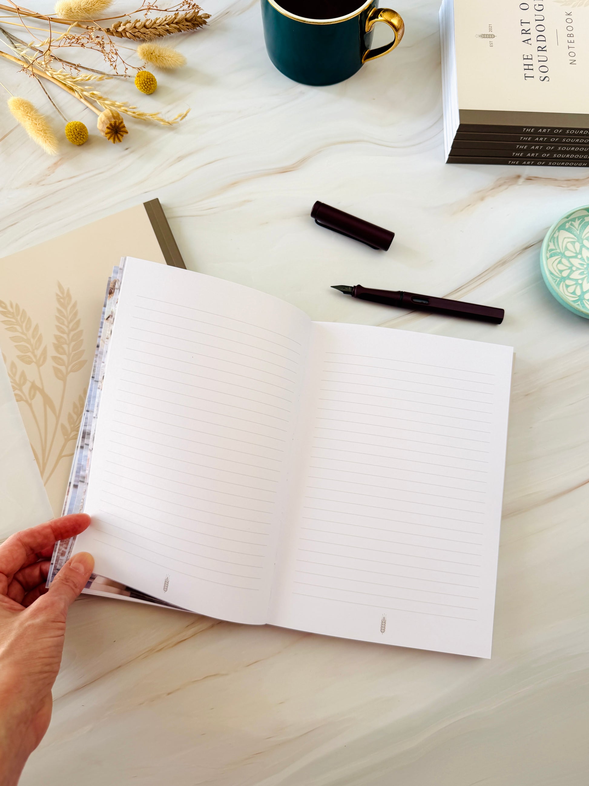 An open lined notebook with a hand holding the left page, placed on a light marble-like surface with a pen, cap, mug, dried flowers, and a book nearby.