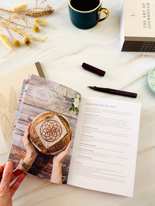 An open printed booklet or recipe book is held in a hand, showing a photo of stencilled sourdough bread on the left page and step-by-step text on the right page. The scene is styled on a light marble-like surface with a dark mug, dried flowers, a pen, and a book titled 'The Art of Sourdough' nearby.